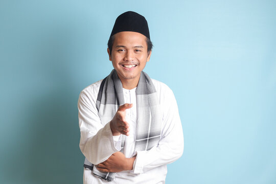 Portrait of attractive Asian muslim man in white shirt reaching out his hand for a handshake, welcoming someone. Isolated image on blue background