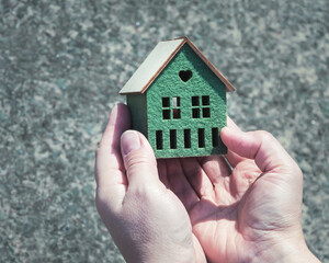 A small house model in female hands on the gray background, selective focus.