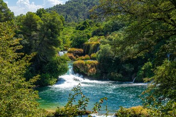 Naklejka na ściany i meble Scenic view of a waterfall flowing through green trees in Krka national park, Croatia on a sunny day