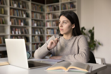 Student girl sit at desk with textbooks and laptop, looks away, thinks about correct answer, search...