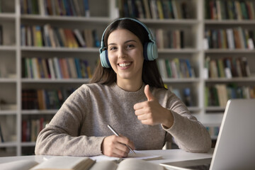 Pretty student 17s girl showing thumb up seated at desk in library, enjoy effective on-line studies...