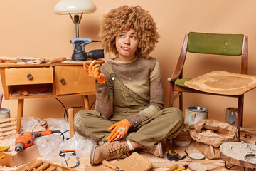 Fototapeta na wymiar Consfused curly woman prepares materials for producing wooden furniture processes wood wears workwear shrugs shoulders does some carpentry work has doubtful expression poses in lotus pose on floor