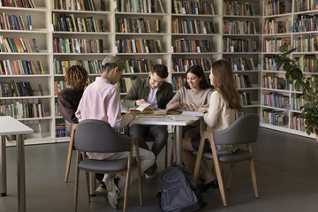 In cozy college library five multi ethnic students, girls and guys engaged in homework making,...