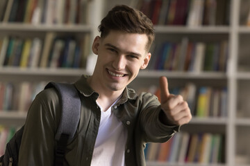 Handsome student guy with backpack pose in college library showing thumbs up to camera, feels...