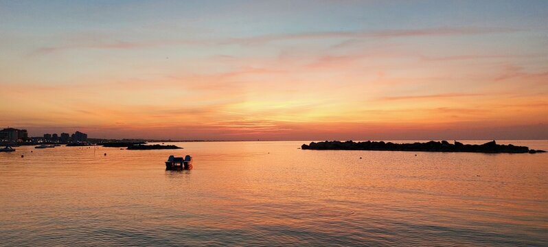 Tramonto al mare, il riflesso del sole da al mare e al cielo sfumature rosse, arancio, rosa e giallo.
Il mare &egrave; calmo, tra gli scogli c'&egrave; una piccola imbarcazione, intorno calma e serenit&agrave;.
