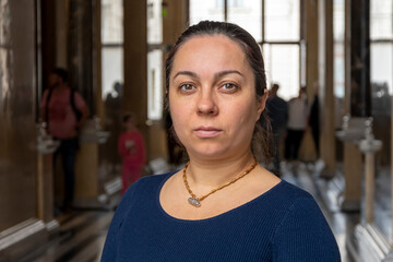 Portrait of a dark-haired adult woman 40-45 years old in a public building, indoors, against the background of windows and people passing by.