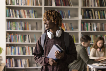 Young African student guy pose in library with schoolmates on background, smile look aside feels...