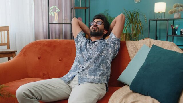 Tired indian man lying down on comfortable couch, taking a rest, daytime nap. Portrait of carefree hindu guy holding hands behind head relaxing, falling asleep at home apartment living room on sofa