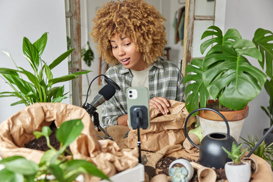 Indoor shot of curly haired young woman films plant care tutorial at home speaks in microphone vlogs on mobile phone at home surrounded by potted flowers. Online streaming and blogging concept
