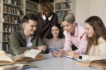 Fototapeta na wymiar Group of attractive teenage multi ethnic schoolmates watch videos, enjoy new cool amusing mobile application gathered in library, distracted from studies using modern smart phone. Generation Z, fun