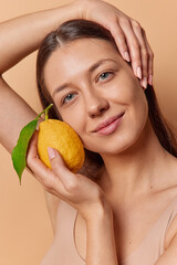 Beauty portrait of dark haired female model leans head holds juicy fresh lemon has natural makeup gets vitamin C for skin care stands against brown background. Wellness and health care concept