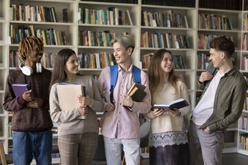Five multi ethnic schoolmates stand in library and talking, having good friendly conversation, wait for lecture start, discuss common task looking satisfied. Education, friendship of diverse teenagers