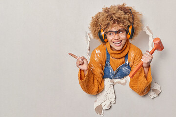 Positive handywoman holds hammer points aside on copy space shows place for your advertisement repairs wall in house wears headphones transparent eyeglasses orange jumper being best repairer ever