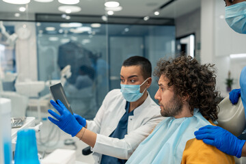 Focused handsome man sitting on dentist's chair watching digital presentation on digital tablet device with two female dental professionals, dentist and her colleague at modern dental clinic.