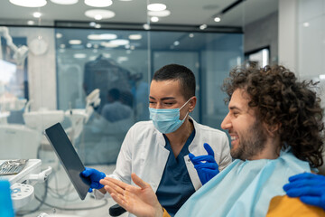 Friendly female dentist with protective face mask using digital tablet showing entertaining online...