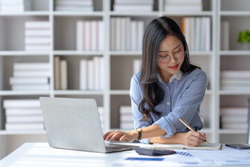 Happy young Asian businesswoman sitting at desk and take notes with laptop computer in the office.