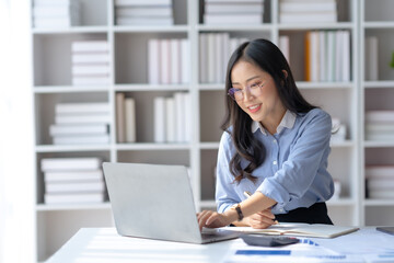 Happy young Asian businesswoman sitting at desk and take notes with laptop computer in the office.