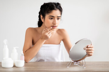 Upset young indian woman touching lips in front of mirror, suffering herpes
