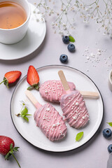 Cake pops ice cream on a stick in pink glaze on a plate with berries on a light background with cup of tea. Healthy homemade sweets.