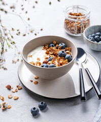 The concept of a healthy breakfast of yogurt, granola and fresh blueberry on a light background with flowers.