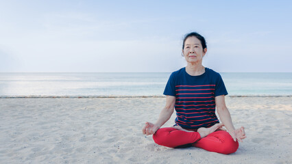 Strong Asian senior elder woman practicing yoga on the beach in summer morning with beautiful blue sky. Healthy lifestyle and fitness leisure concept.