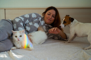 Caucasian woman holding white fluffy cat and Jack Russell Terrier dog lying on bed. Redhead girl hugging pets. 