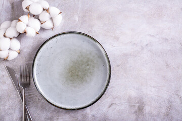 Empty ceramic plate, cutlery and cotton branch on gray background top view