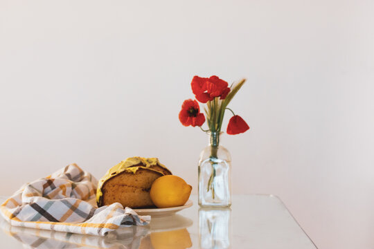 Rural still life with red poppies in glass vase and a plate with biscocho Bizcocho lemon muffin on kitchen table. Sweet dessert prepared at home. A red cup of coffee or tea. Biscotto pastries, cakes.