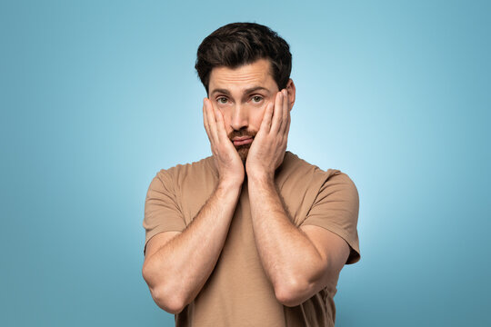 Frustrated unhappy man keeping hands on cheeks, feeling tired and exhausted reluctant to do something, blue background