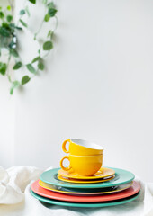 Empty yellow cups and brightly colored plates on a white table covered with a linen tablecloth, a flower pot on the wall in the background. Vertical photo.