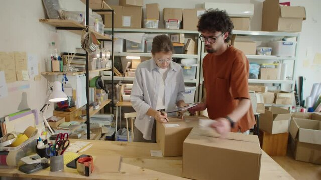 Woman using digital tablet as male coworker sticking address labels to cardboard boxes while preparing online orders for shipping in delivery service office