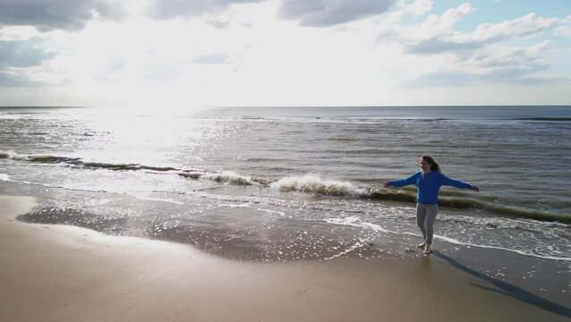 Young girl on sandy coast. Girl leaves footprints on sand. Attractive woman in blue sweater on beach. Girl walking along seashore. Aerial view of seashore. Beautiful ocean waves. Beach with  sand