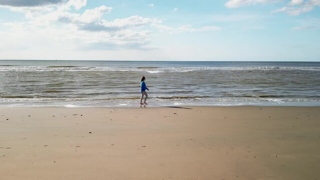 Young girl on sandy coast. Girl leaves footprints on sand. Attractive woman in blue sweater on beach. Girl walking along seashore. Aerial view of seashore. Beautiful ocean waves. Beach with  sand