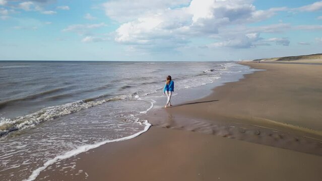 Young girl on sandy coast. Girl leaves footprints on sand. Attractive woman in blue sweater on beach. Girl walking along seashore. Aerial view of seashore. Beautiful ocean waves. Beach with  sand