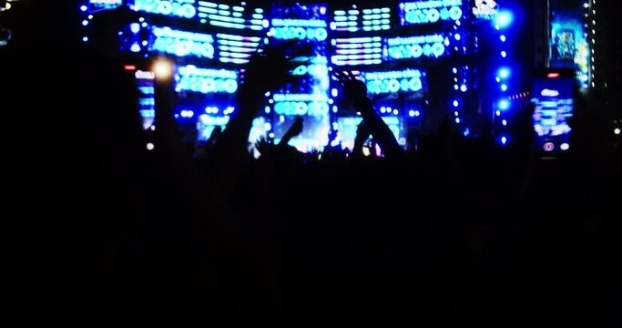 Pop band concert on street at night. Silhouettes fans raise their hands in front bright colorful strobe lights on stage.Crowd is dancing, filming performance rock band on their phones, throwing party.