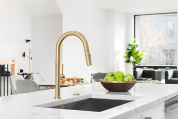 A gold kitchen faucet detail with a white marble countertop, white cabinets, and decorations in the...