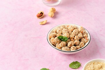 Sweets, peanuts in honey and sesame seeds in a white ceramic bowl on a pink concrete background. Useful sweets, handmade candies.