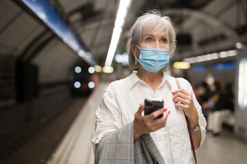 Mature European woman in mask waiting for subway train in metro station and using her smartphone.
