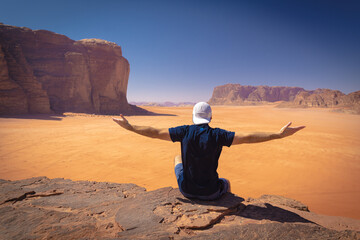 Wadi Rum w Jordanii. Mężczyzna siedzący na pustynnej skale patrzący w dal. - obrazy, fototapety, plakaty