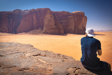 Wadi Rum w Jordanii. Mężczyzna siedzący na pustynnej skale patrzący na pustynię.  - obrazy, fototapety, plakaty