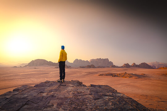 Wadi Rum w Jordanii. Mężczyzna stojący na pustynnej formacji skalnej patrzący na wsch&oacute;d słońca. 