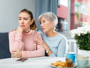 Senior woman calming chagrined young adult daughter sitting at table at home .
