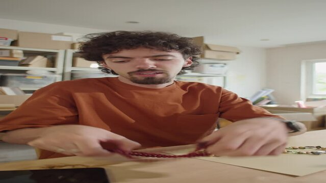 Vertical POV shot of young man demonstrating handmade beaded jewelry and talking on camera while filming DIY blog or video advertising for online store