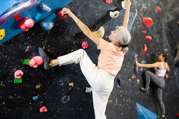 Positive focused mature woman exercising on black climbing wall without safety belts and preparing for summer mountain hiking in game center indoors