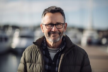 Portrait of a handsome middle-aged man with gray hair and beard wearing eyeglasses standing in front of a yacht marina