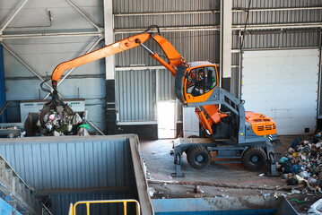 Grabbing excavator near trash heap in warehouse of plant