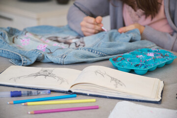Unrecognizable woman painting a design of a deer and flowers in a denim jacket. Woman's creative expression through painted clothing designs.