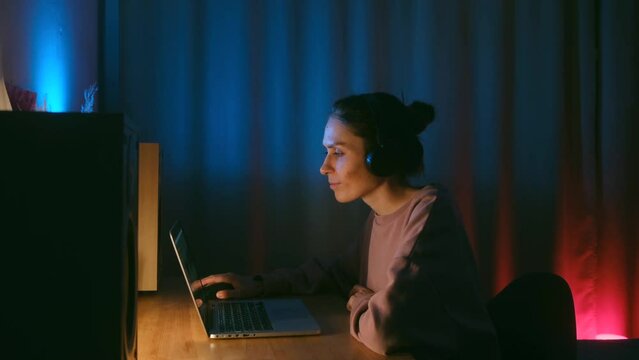 Cute person at night at a laptop with headphones. Neon light, a young woman composing music at home, dancing to the beat. High quality 4k footage