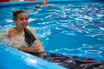 Young woman in a bright swimsuit swims in the pool with a dolphin. An enthusiastic woman shows her tongue to the camera in pleasure, holding on to her dorsal fin.