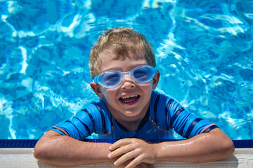 Smiling boy poses for the camera emerging from the water in a summer pool wearing swimming goggles....